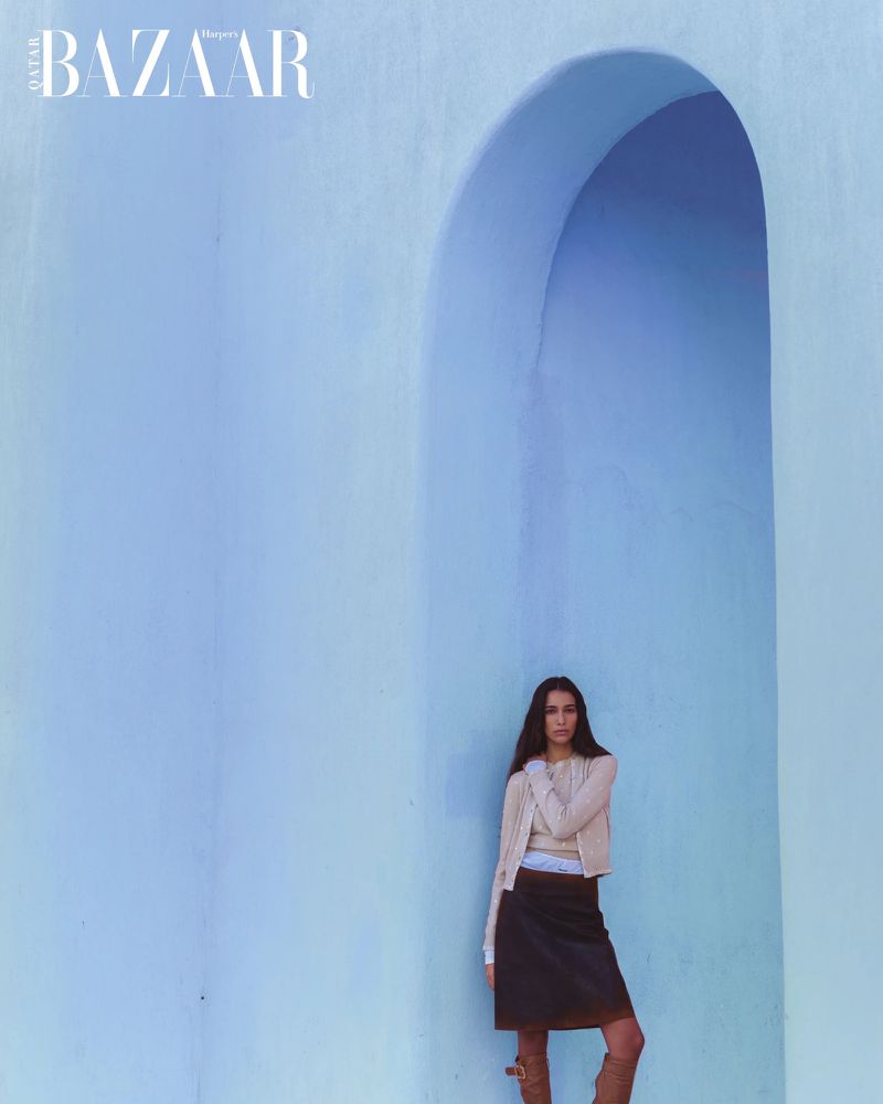 A woman in a skirt and cardigan poses in front of a blue wall with an arched opening for a fashion editorial magazine shoot.