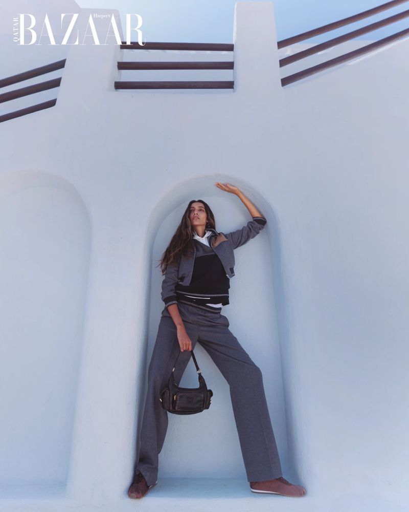 Fashion shot of a woman in a grey jacket and pants holding a purse, posing in front of a white wall with blue sky at the top.