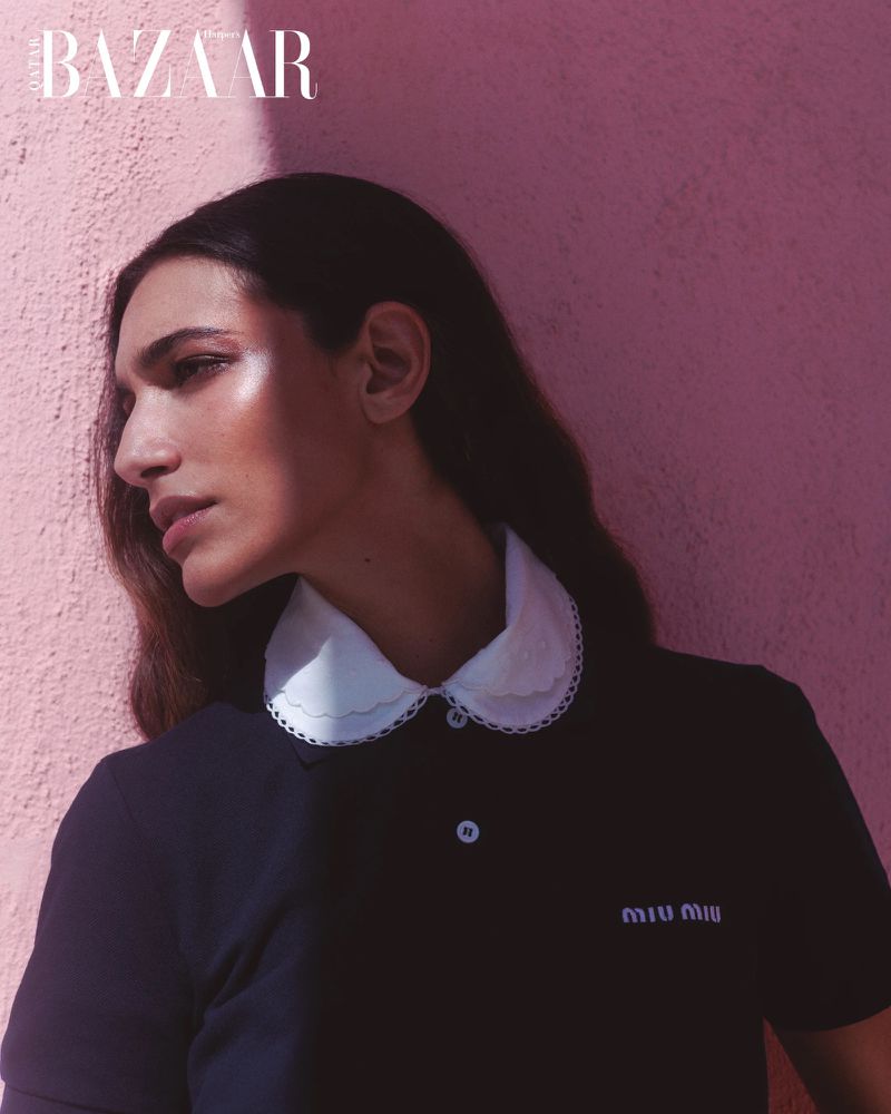 Portrait of a woman wearing a navy polo with a white collar against a pink wall. The Bazaar logo is in the upper left corner.