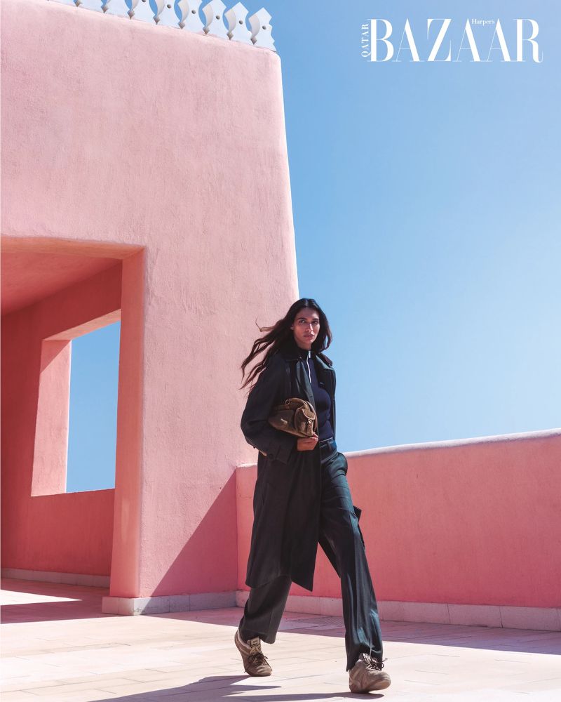 Fashion editorial shot of a woman in a black coat carrying a bag, posing on a pink rooftop under a bright blue sky, Harpers Bazaar logo visible.