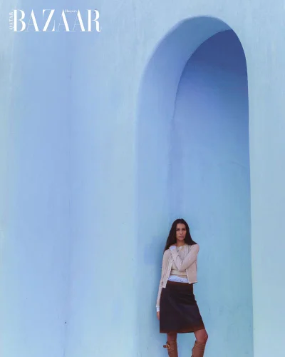 Model Nour Rizk poses in a beige knit cardigan and dark brown skirt framed by a pale blue architectural archway for Harper's Bazaar Qatar.