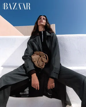 Model Nour Rizk wearing an oversized dark grey coat and tan handbag, posed against white architecture and a clear blue sky.