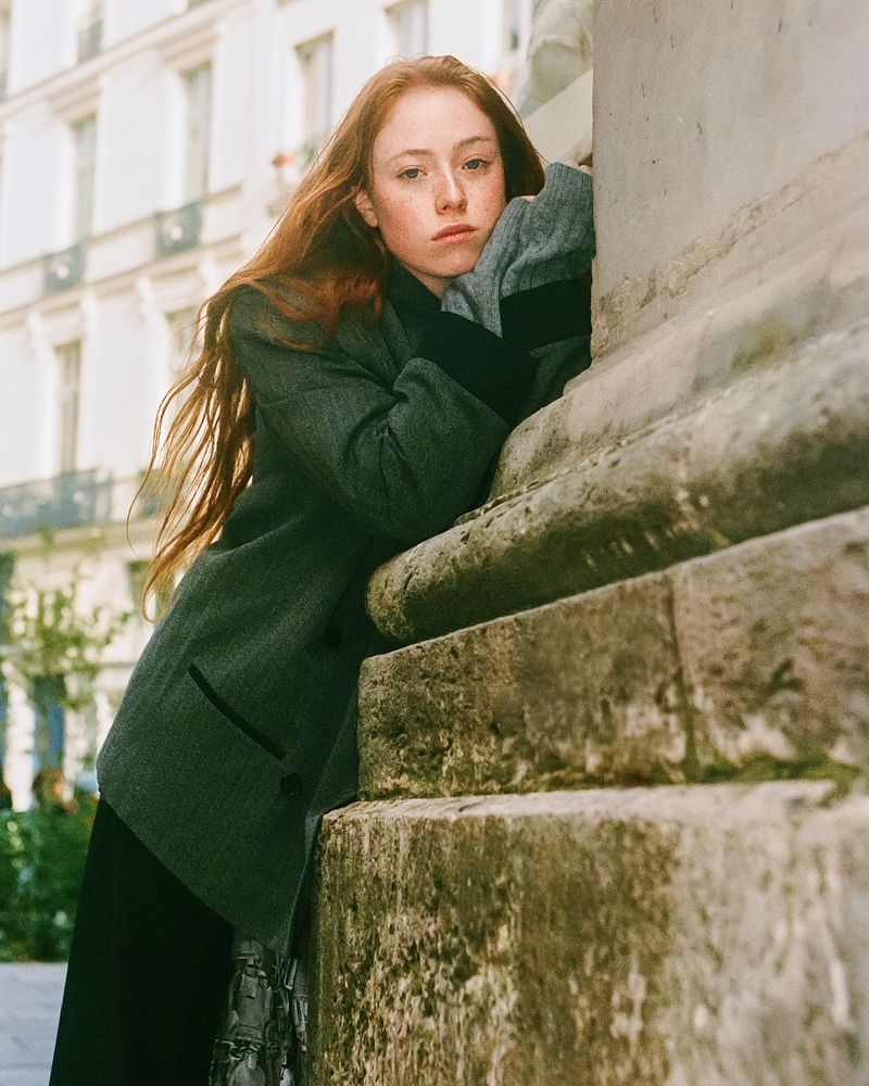 Portrait of a woman with red hair leaning against a stone column outdoors, wearing a grey jacket, with buildings in the background.