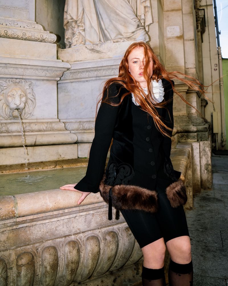 Redhead woman in a black jacket and tulle top stands in front of a stone fountain with a lion's head and flowing water.