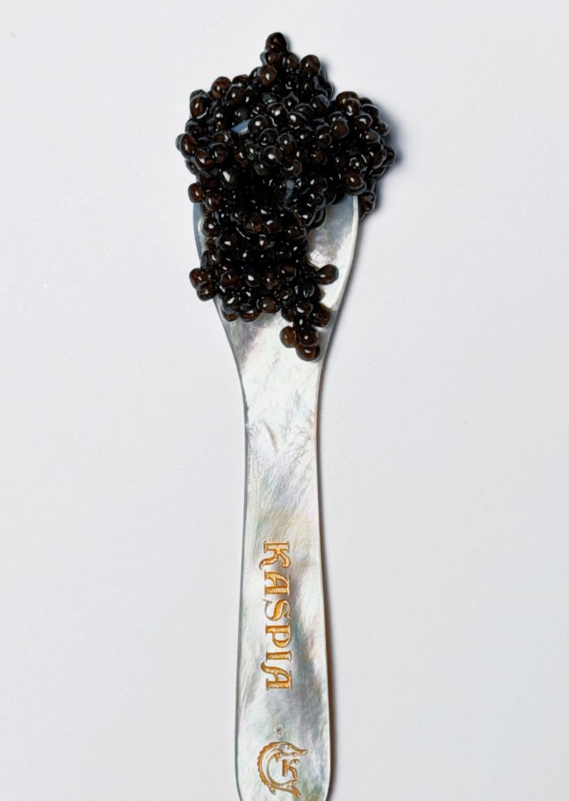Close-up of black caviar piled on a mother-of-pearl spoon with 'Kaspia' inscription, set against a clean white background.