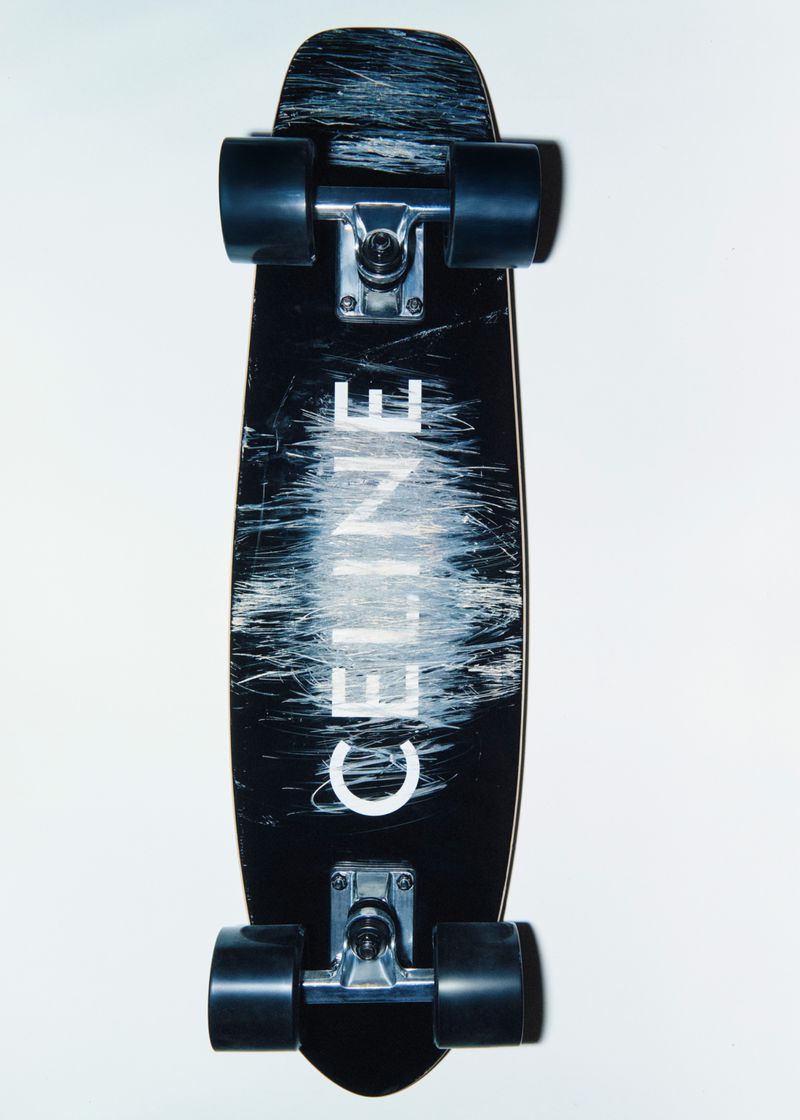 A black skateboard with a white CELINE logo and black wheels on a white background, showcasing its design.