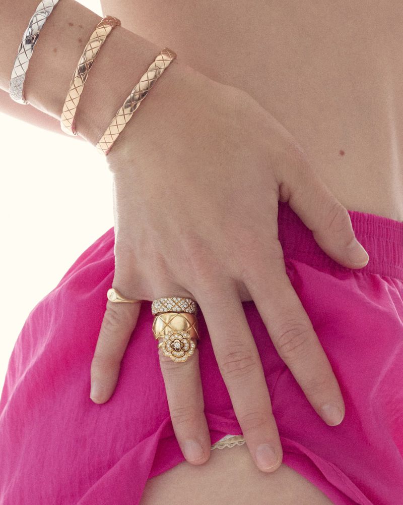 Close-up of a hand with three rings, adorned with silver and gold bracelets, set against a blurred backdrop of pink fabric.