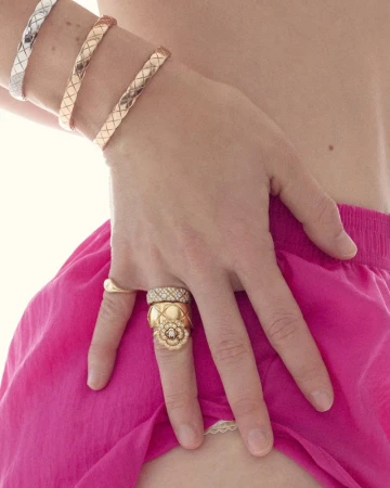 Close-up of a hand with three rings, adorned with silver and gold bracelets, set against a blurred backdrop of pink fabric.