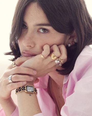 Close-up of an adult woman with brown hair wearing a pink collared shirt, gold rings, gold stud earrings, and a gold watch with a black band.