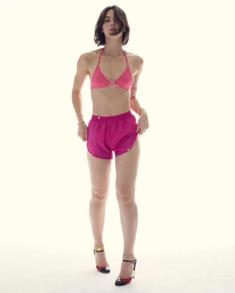 Woman poses in a pink bikini top and shorts against a bright white backdrop, accessorized with heels and jewelry.
