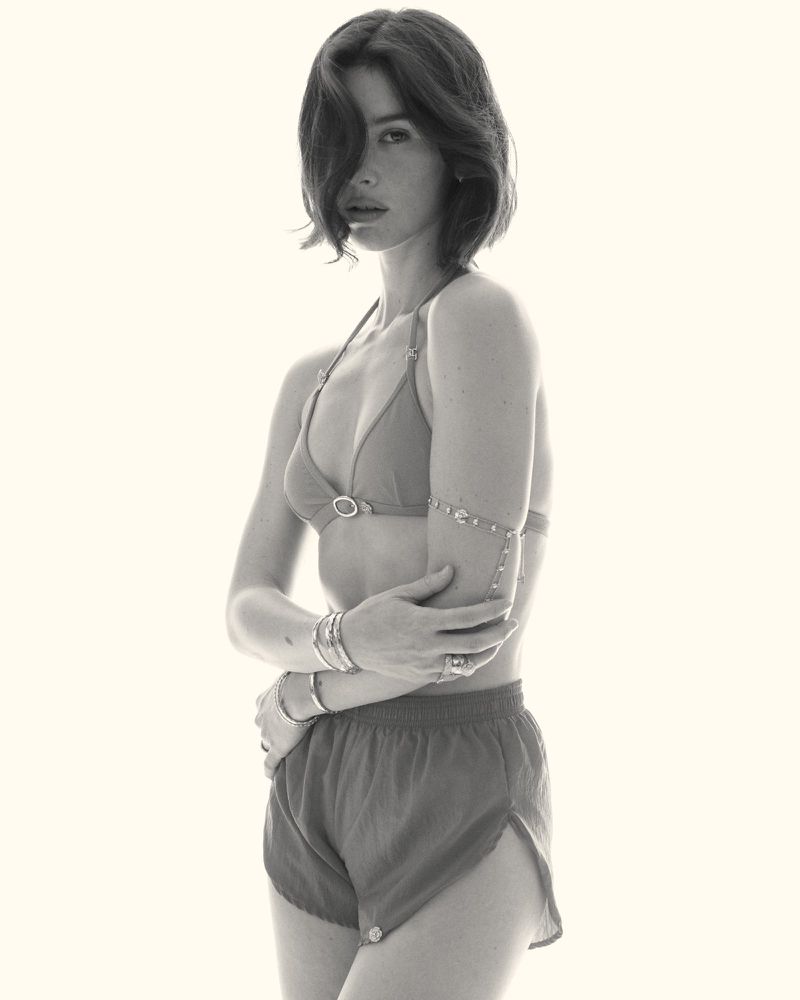 Gracie Abrams wearing a triangle bikini top, shorts, and layered arm jewelry against a plain white studio backdrop.