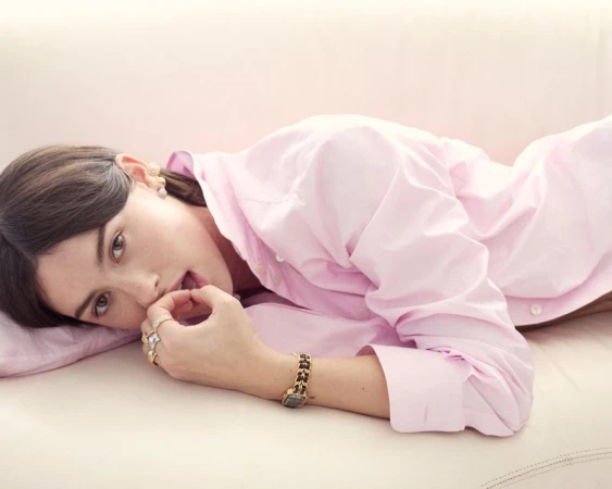Gracie Abrams reclining on a cream couch wearing an oversized pink button-down shirt, gold watch, and rings.