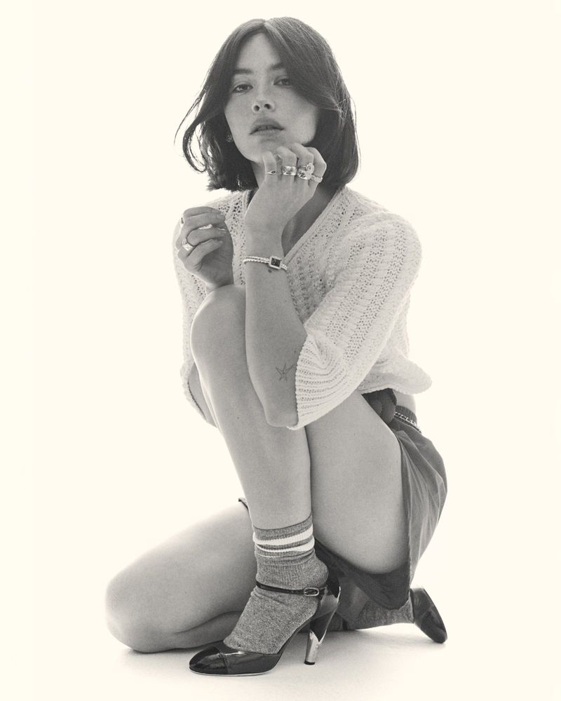 Monochrome portrait of a brunette woman kneeling in a studio, wearing a knit sweater, skirt, socks, and patent leather heels.