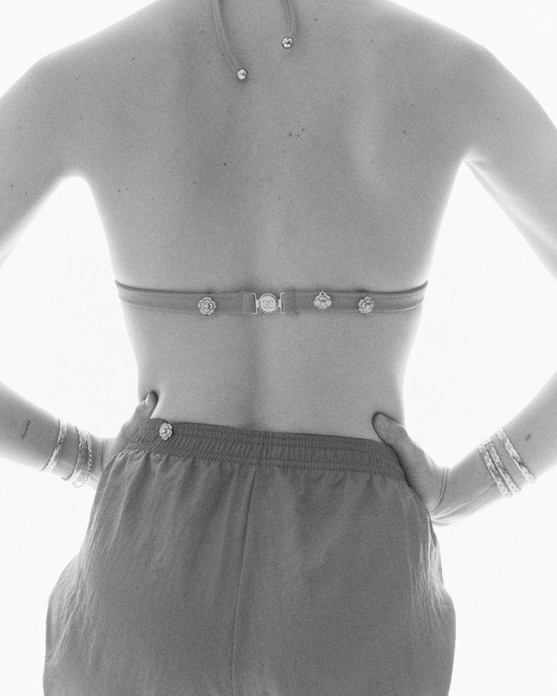 Black and white photo of a slender adult woman in a bikini top and shorts, facing away from the camera, with hands on her hips.