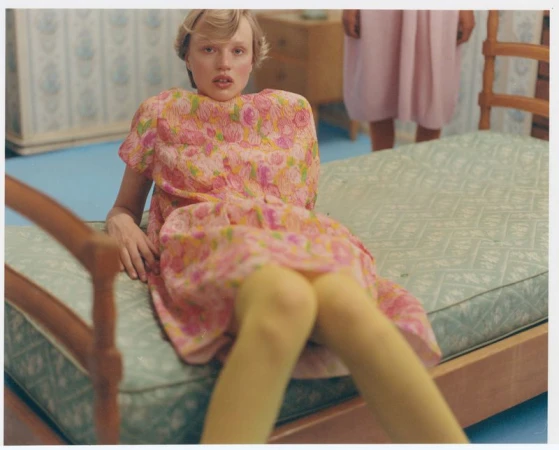 A fair-skinned young adult poses on a vintage daybed in a floral dress and yellow tights, while another figure stands in the background.