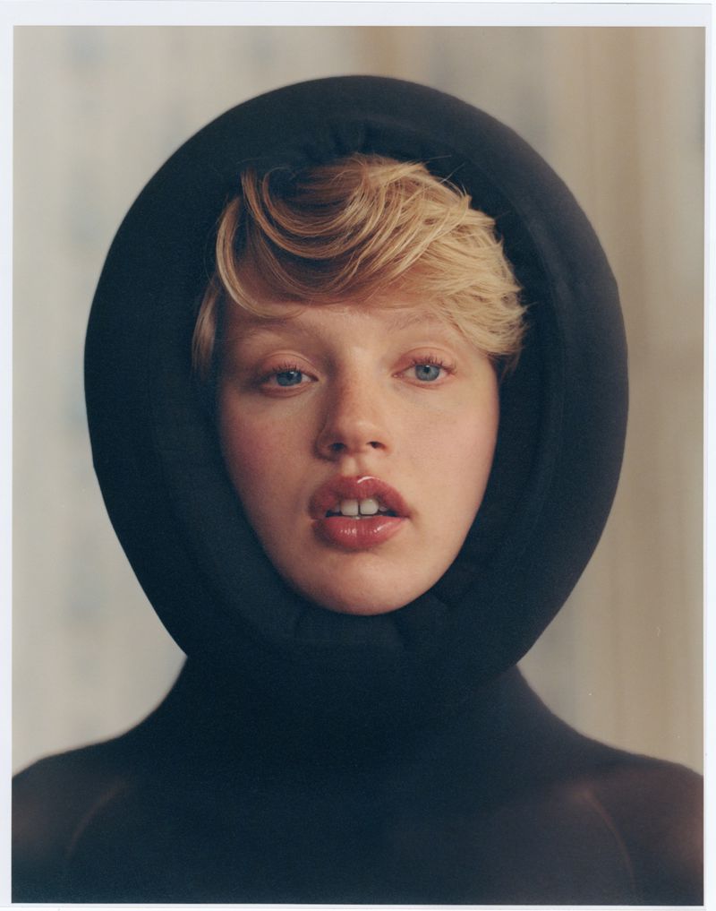 Close-up portrait of a young adult female with blonde hair and glossy lips, wearing a puffy black hood, with a blurred floral backdrop.