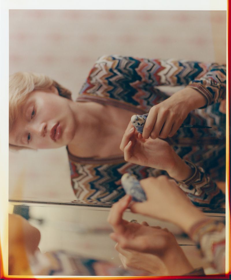 Young fair-skinned woman with short blonde hair applying blue nail polish in front of a mirror with patterned wallpaper in the background.