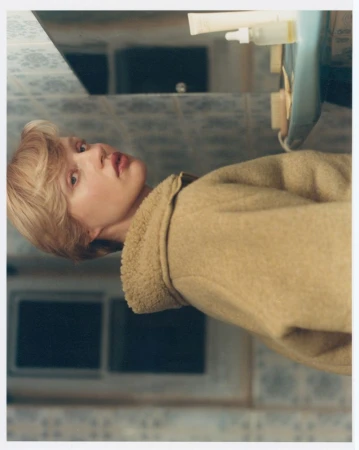 Woman in a tan coat indoors with neutral walls and patterned backsplash, portrait-style capture, looking at camera with soft, diffused lighting.