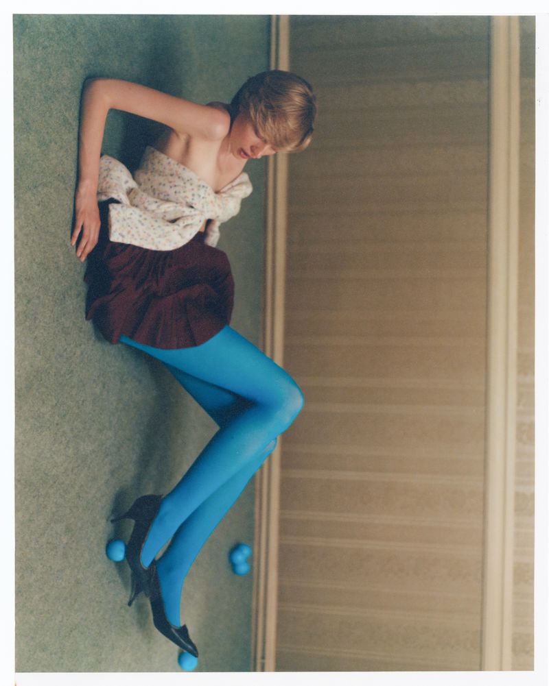 Young woman poses in vintage fashion with blue tights, mini-skirt, and heels in a studio environment with light green carpet.