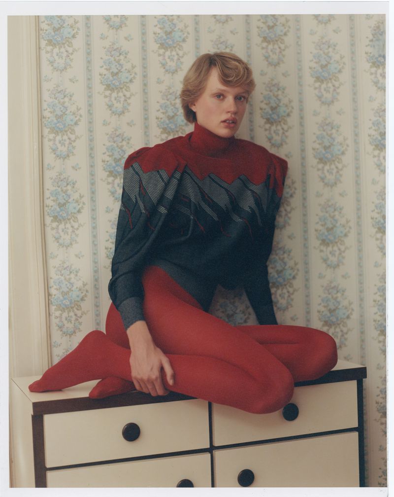 A fair-skinned woman with short blonde hair sits on a white dresser wearing red tights and a dark gray and red sweater in a room with floral wallpaper.