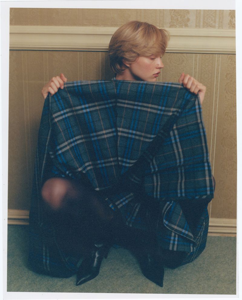Woman sits on a carpeted floor, holding a large blue and grey plaid textile up to her face. Neutral wall is in the background.