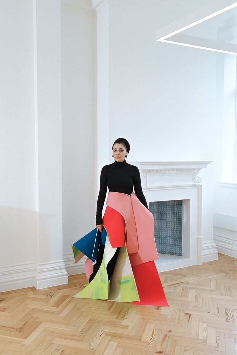 A woman stands in a bright room with a colorful sculptural dress-like garment made of paper or fabric, holding a triangular blue object.