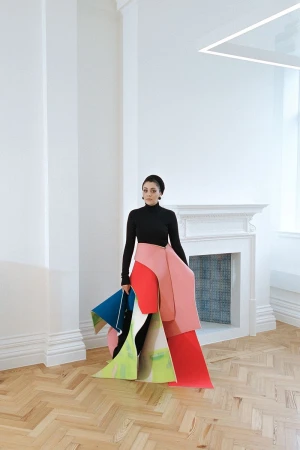 A woman stands in a bright room with a colorful sculptural dress-like garment made of paper or fabric, holding a triangular blue object.