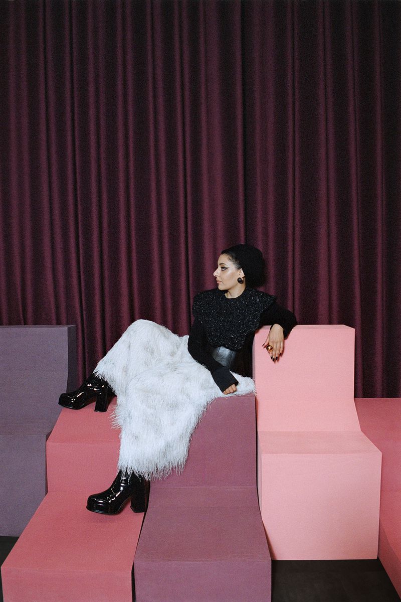 Adult woman in black top, long faux-fur skirt, and black boots sitting on pink block in front of a maroon curtain backdrop.