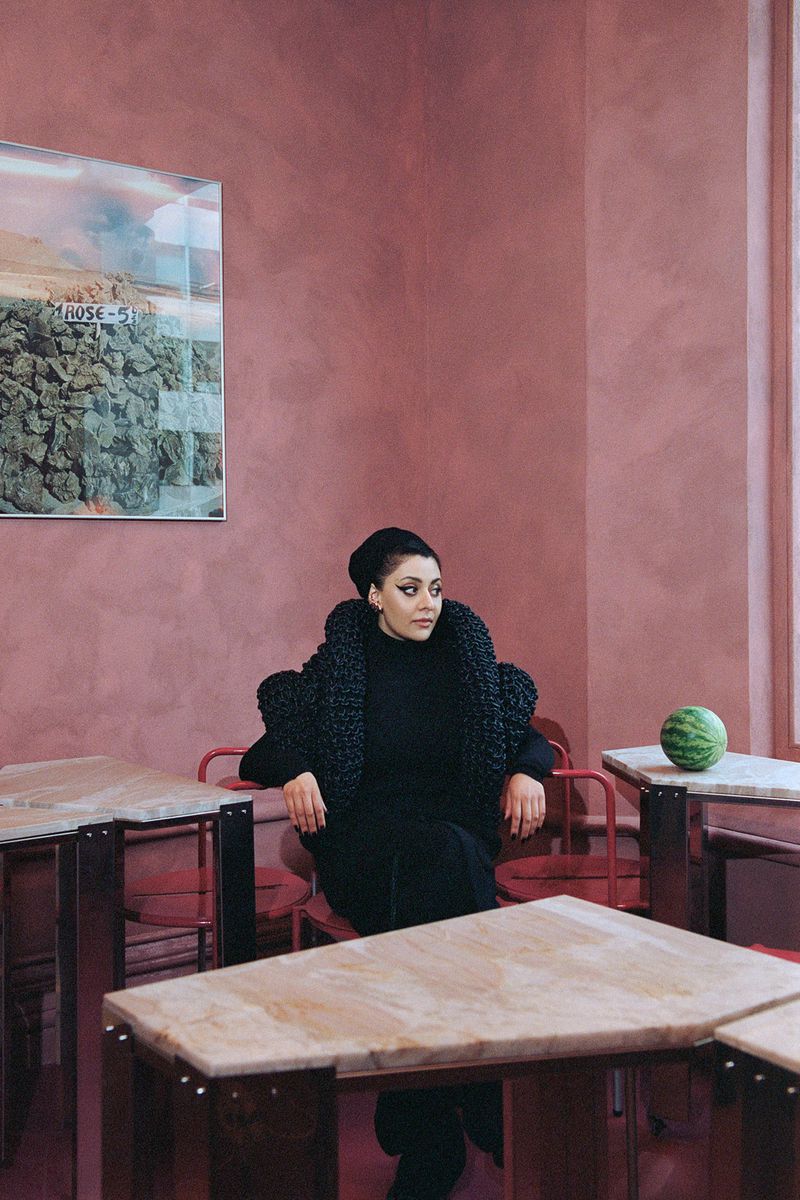 An adult female with dark hair is sitting in an indoor setting near tables and chairs, wearing an all-black outfit and dark patterned jacket.