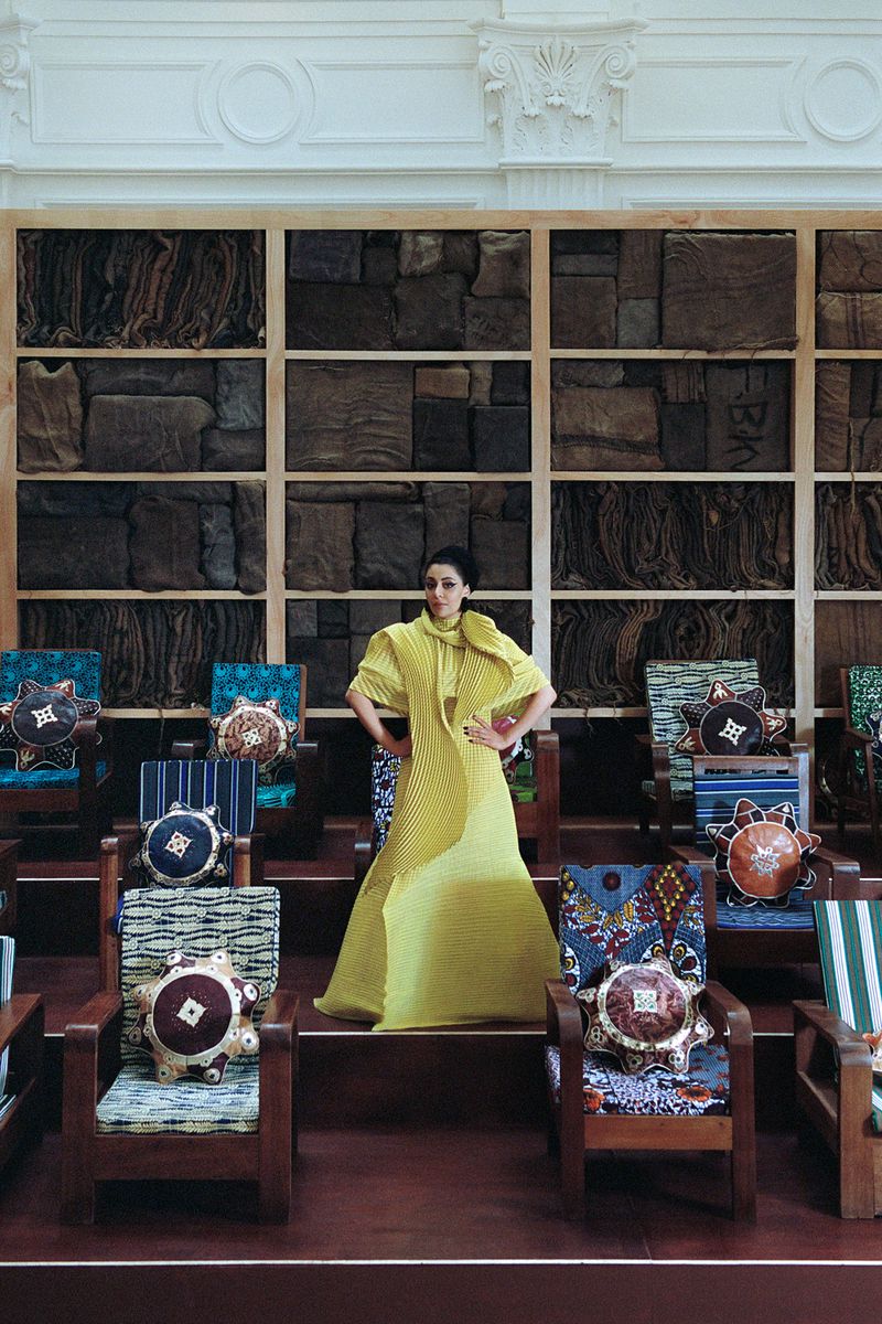 A woman in a yellow dress stands in front of chairs with patterned pillows and a wall filled with fabric swatches.