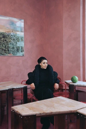 Sumayya Vally seated in a room with pink textured walls, wearing a black structured coat and turtleneck, photographed by Oda Eide for Tank Magazine.