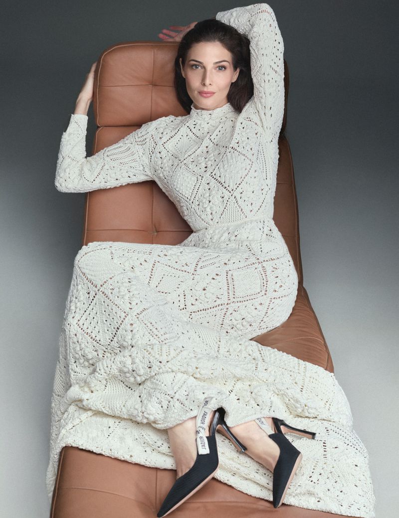 Adult woman poses in a studio setting wearing a white knit dress while sitting in a leather chair. She wears black heels with white accents.