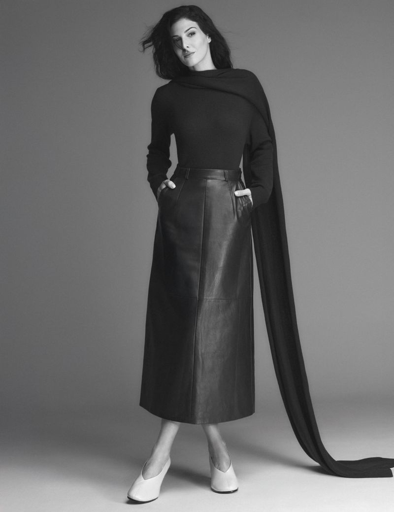 Monochrome portrait of a woman in a black top, leather skirt, draped scarf, and white shoes, standing against a gray background.