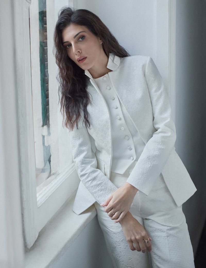 A striking portrait of a woman in an elegant white suit, leaning against a window, exuding sophistication and style in an indoor setting.
