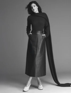 Razane Jammal posing in a black long-sleeve top, floor-length scarf, and black leather midi skirt against a grey studio backdrop.