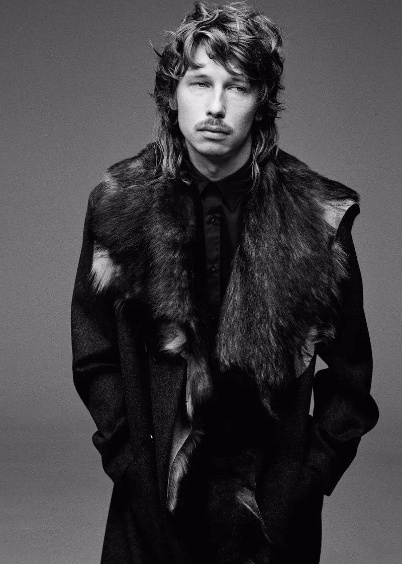A man with a mullet and mustache poses indoors wearing a dark coat with a fur collar, showcasing a black and white fashion portrait.
