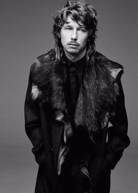 A man with a mullet and mustache poses indoors wearing a dark coat with a fur collar, showcasing a black and white fashion portrait.