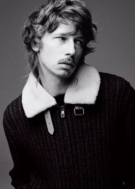 Monochrome portrait of a stylish adult male with wavy hair, wearing a wool zip jacket with a white fur collar in a neutral studio setting.