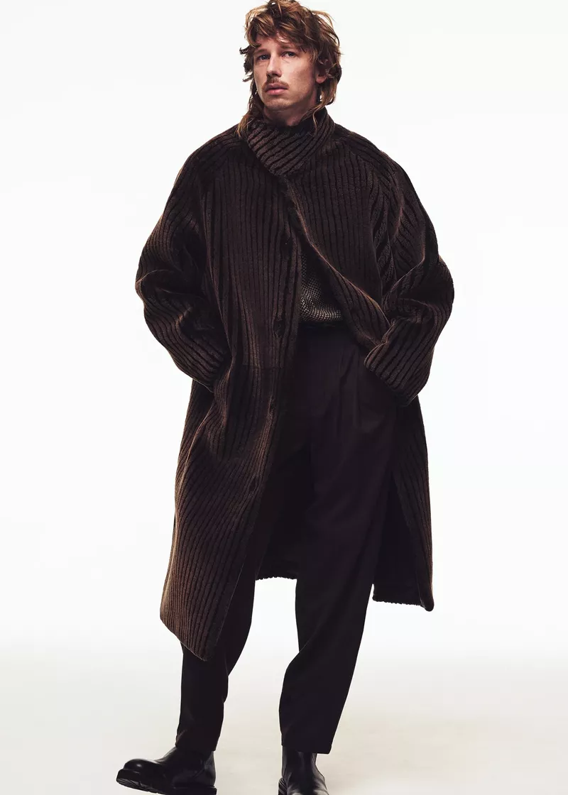 A male model poses in a full-length shot wearing a brown coat and dark pants against a white backdrop, showcasing a relaxed fashion style.