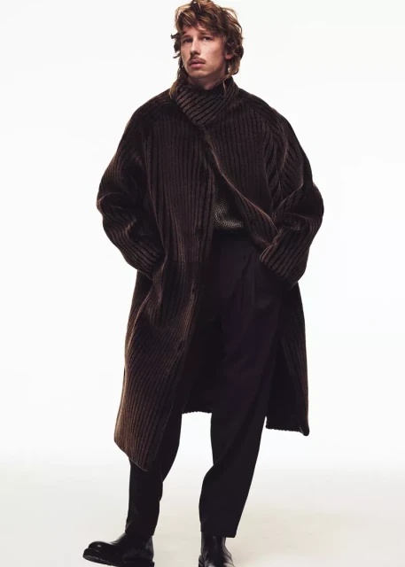 A male model poses in a full-length shot wearing a brown coat and dark pants against a white backdrop, showcasing a relaxed fashion style.