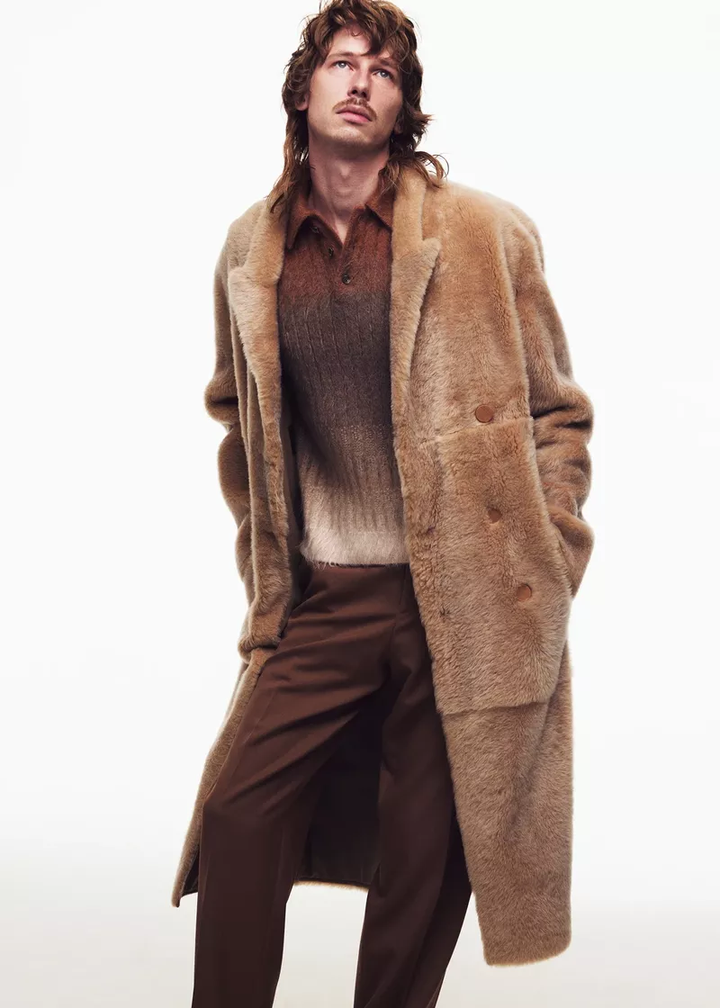Fashionable adult male poses in a tan shearling coat, brown polo shirt, and trousers against a bright white backdrop in a fashion editorial.