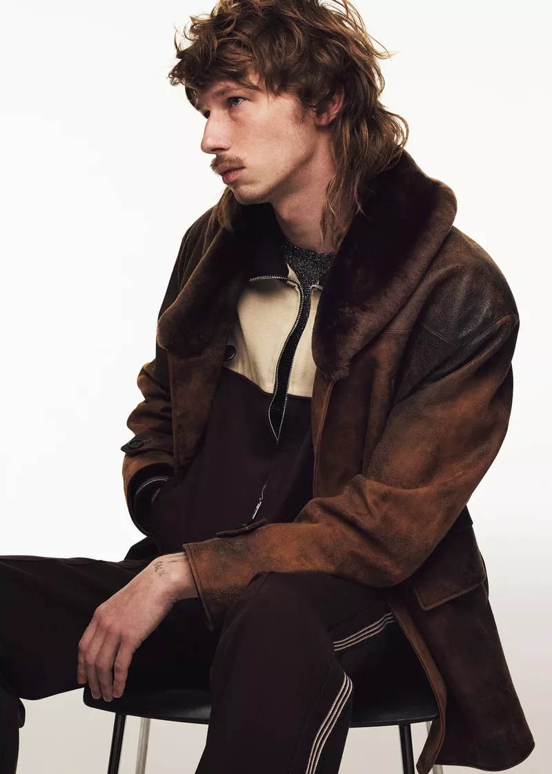 Profile shot of a man with a mullet, wearing a brown jacket and sitting on a black chair against a bright white backdrop.