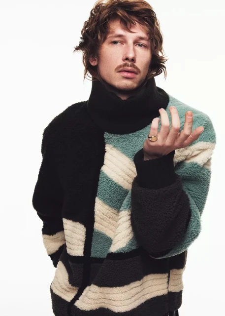 Man with brown hair poses in a studio wearing a geometrically patterned fleece turtleneck sweater against a clean white backdrop.