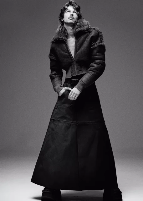Black and white portrait of a male model wearing a fur-collared jacket and long skirt looking up in a photo studio setting. Fashion shoot.