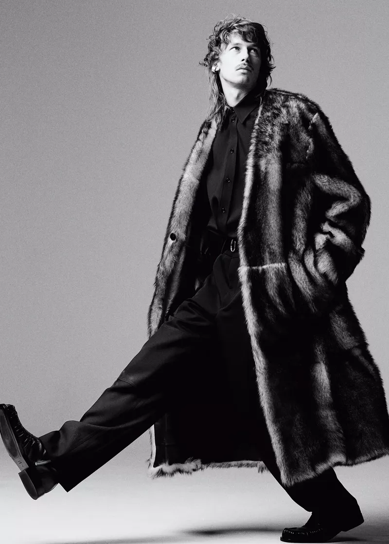 Model Matïss Rucko strides across a studio set wearing a long fur coat, black button-up shirt, trousers, and dark boots.
