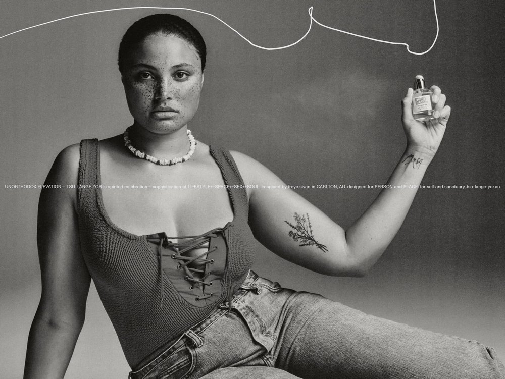 Adult female poses in front of grey background, wearing a low-cut top and jeans while holding perfume. Focus on fashion and beauty.