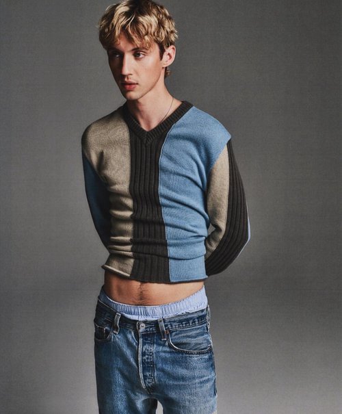 Portrait of a young, blonde-haired man wearing a color-blocked sweater and jeans, looking off-camera against a simple gray background.