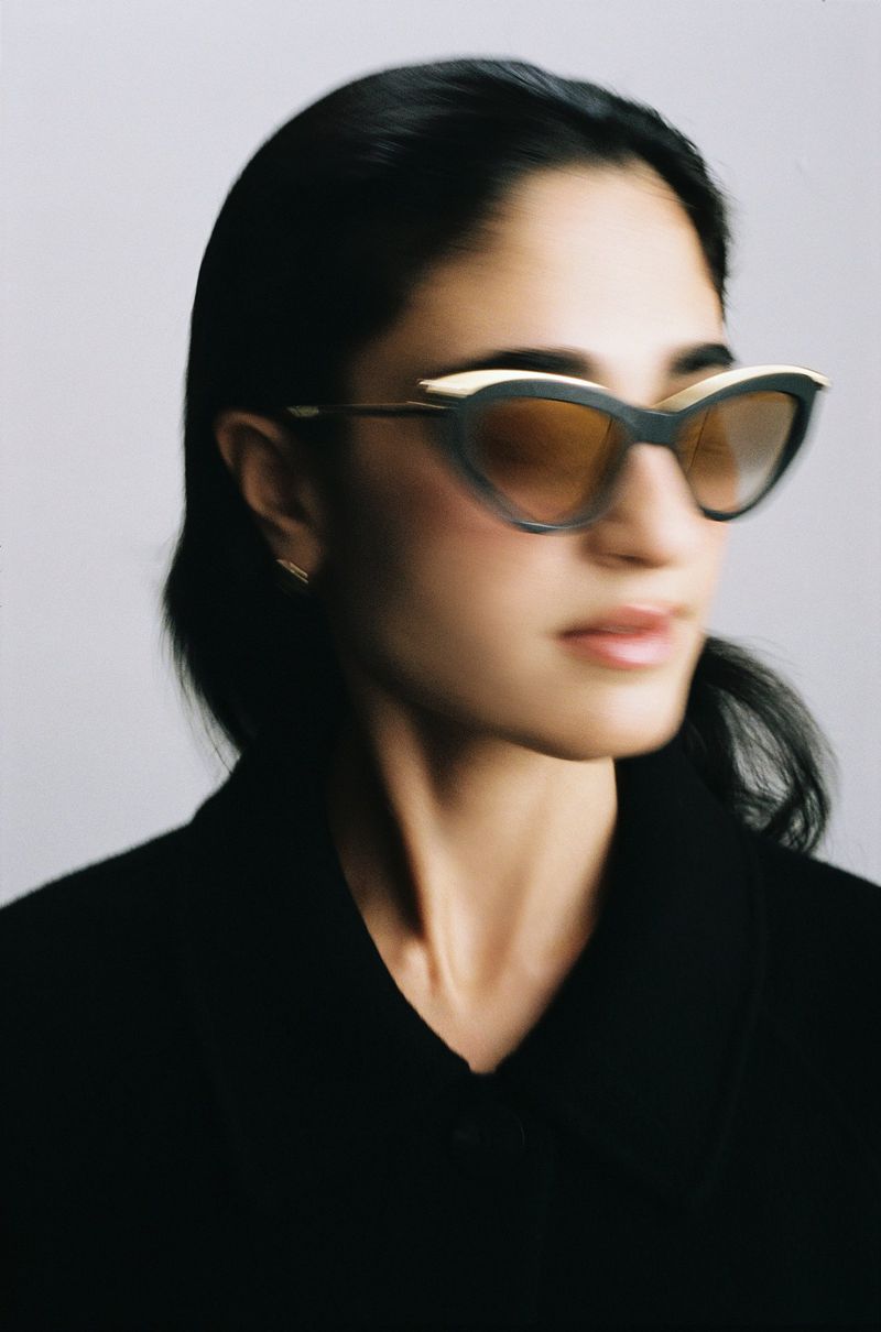 Headshot of an adult woman with black hair wearing black-and-gold cat-eye sunglasses and a black jacket against a white background.