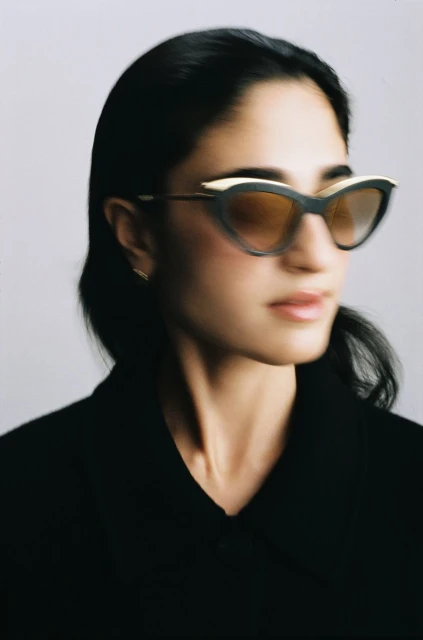 Headshot of an adult woman with black hair wearing black-and-gold cat-eye sunglasses and a black jacket against a white background.