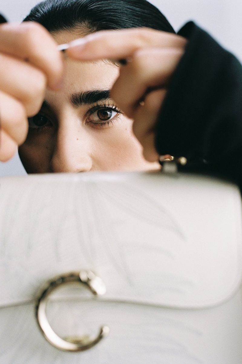 Woman with dark hair gazes at camera over white marble-patterned purse with gold C-shaped clasp, fingers framing face, neutral backdrop.